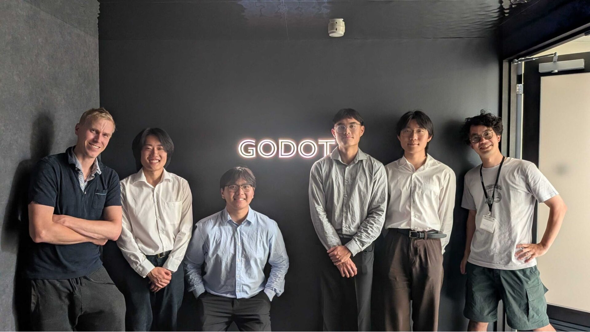 壁面のGODOTロゴを背景に並んで立つ6名の人物の集合写真。