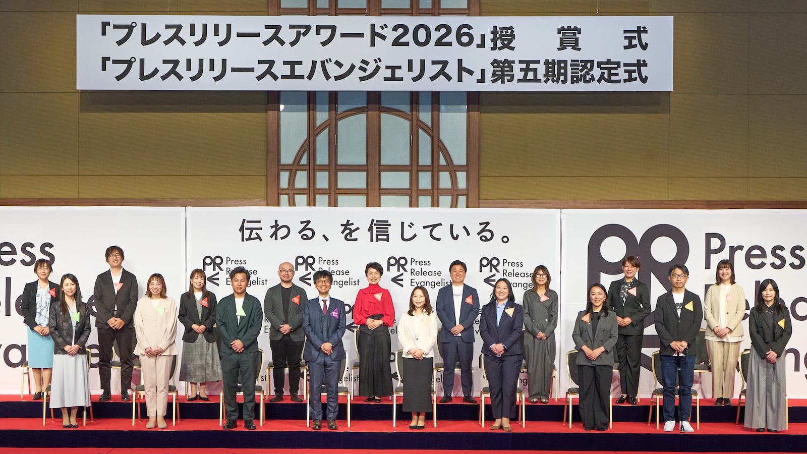 認定式の看板の前で、壇上に一列に並んで立つ参加者の集合写真