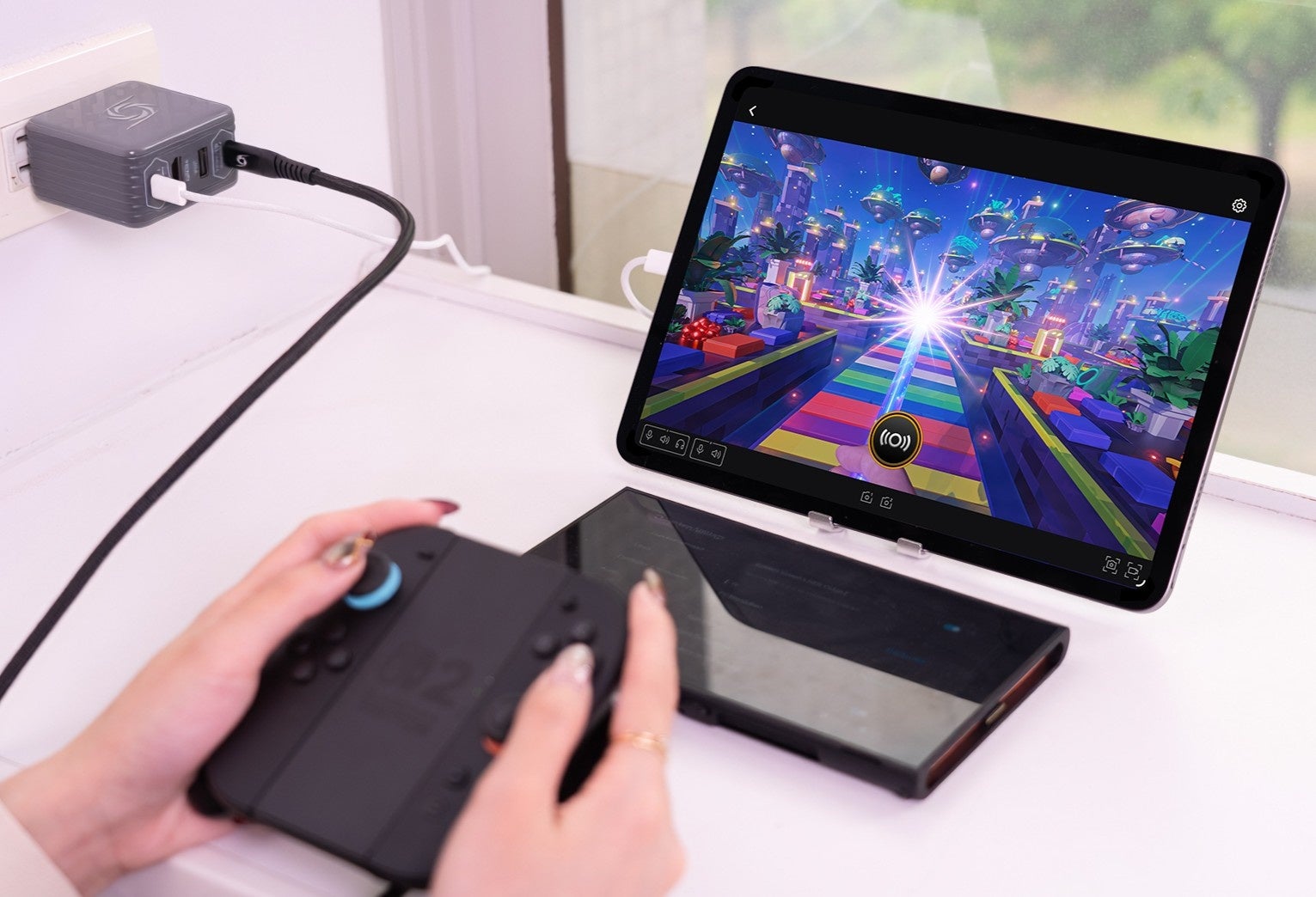 壁面コンセントに接続された充電器型機器とタブレット画面、ゲーム機コントローラーを操作する手元の様子
