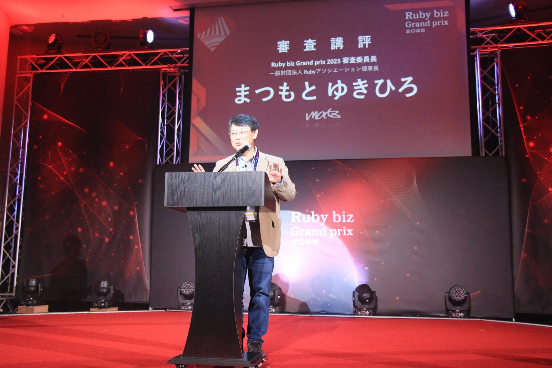 演台に立ちマイクを通して講評を行う人物の姿と背後のスライド