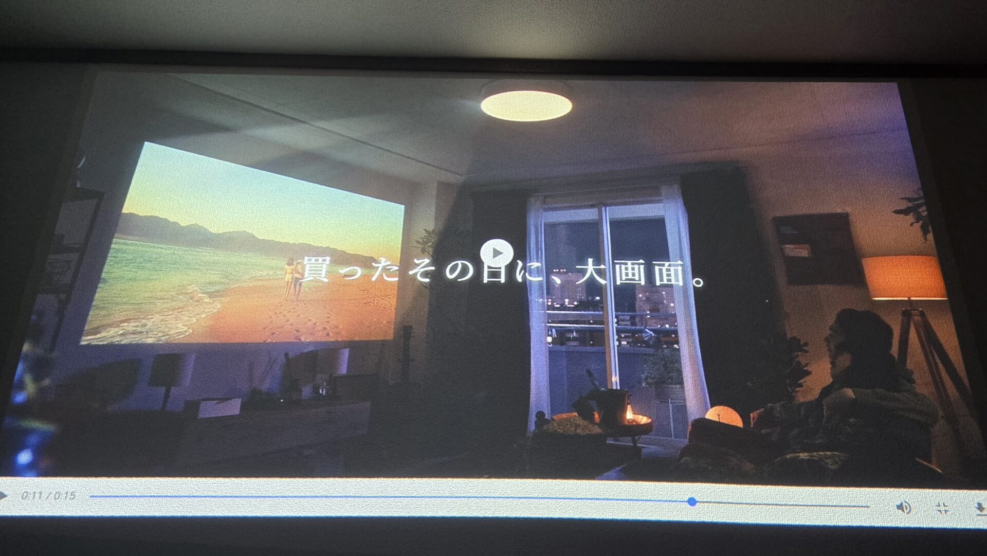 プロジェクターで壁に映された砂浜と海の映像に「買ったその日に大画面」の文字ソファに座る人のいる部屋の様子