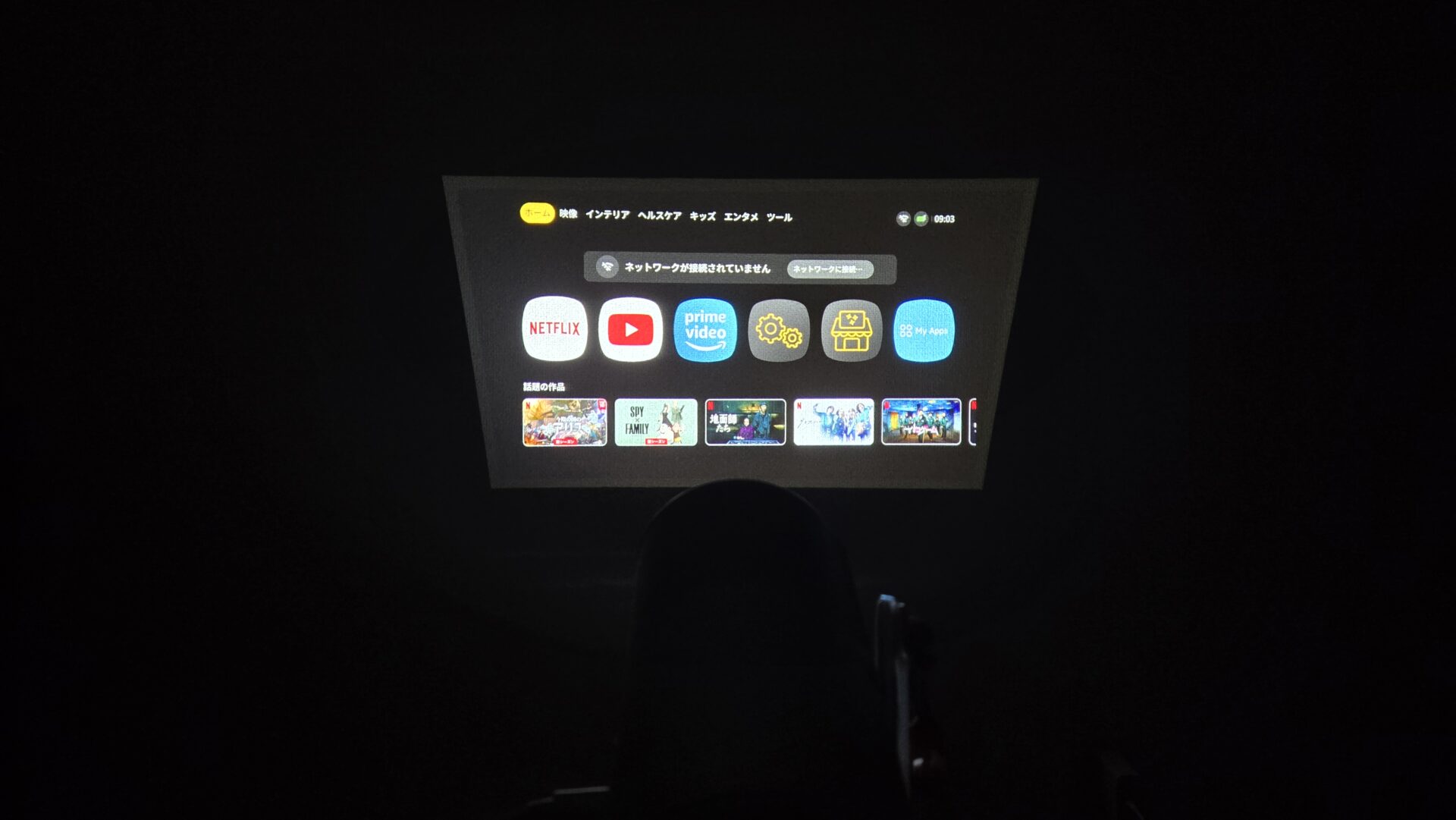 暗い部屋でプロジェクターが壁に映し出すホーム画面 ネット未接続の表示とNetflix YouTube Prime Videoなどアプリのアイコン アニメ作品のサムネイルが見える