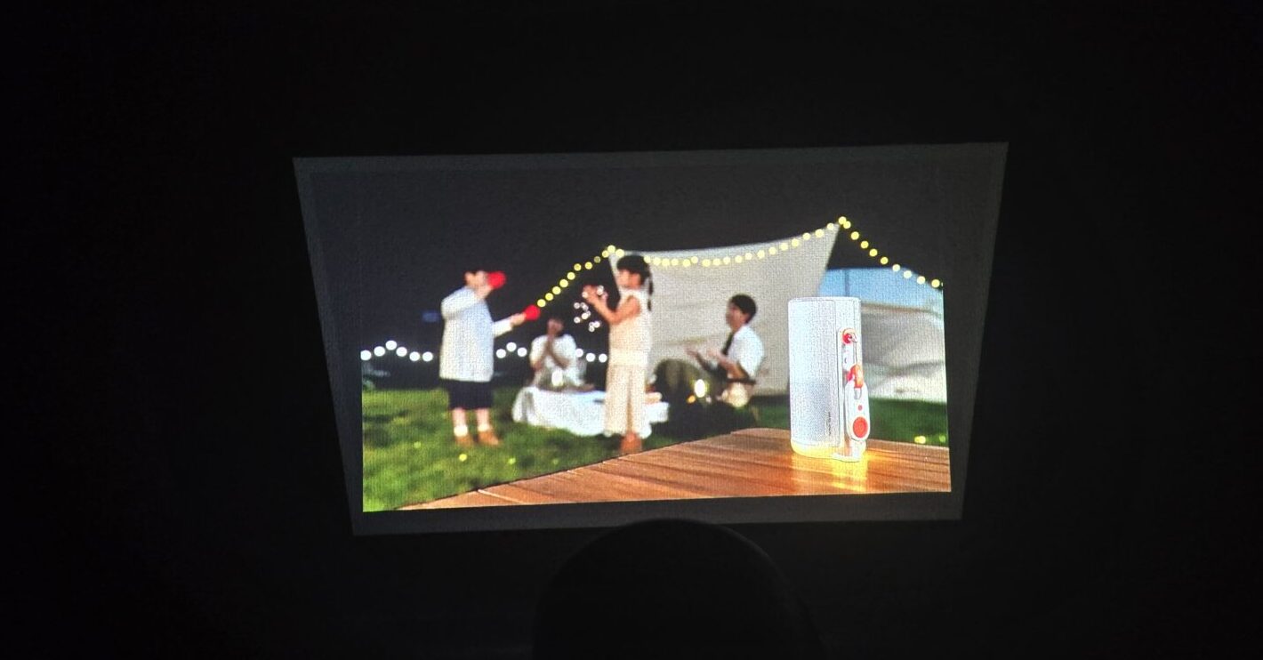 暗い部屋で白い縦長の機器がテーブルに置かれ壁には夜のキャンプ風景が投影されている