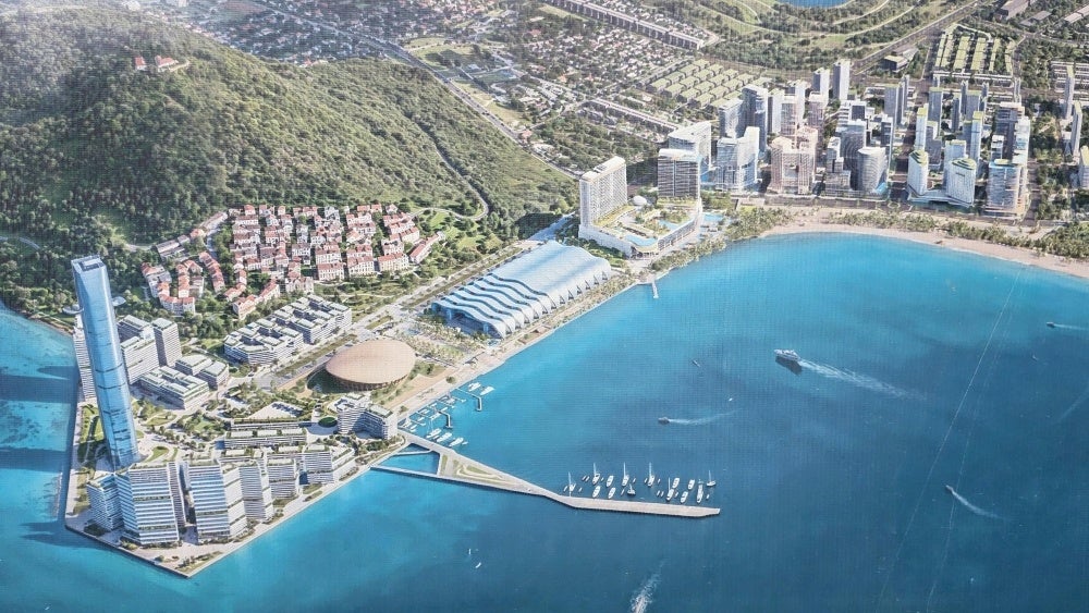青い海に面した沿岸部に並ぶ高層建築物やスタジアム形状の施設、マリーナを描いた開発エリア全体の鳥瞰パース図。