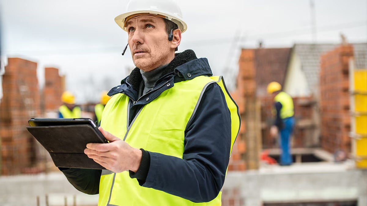 ヘルメットとベストを着用した人物が作業現場でタブレットを持ち、ヘッドセットを装着している写真