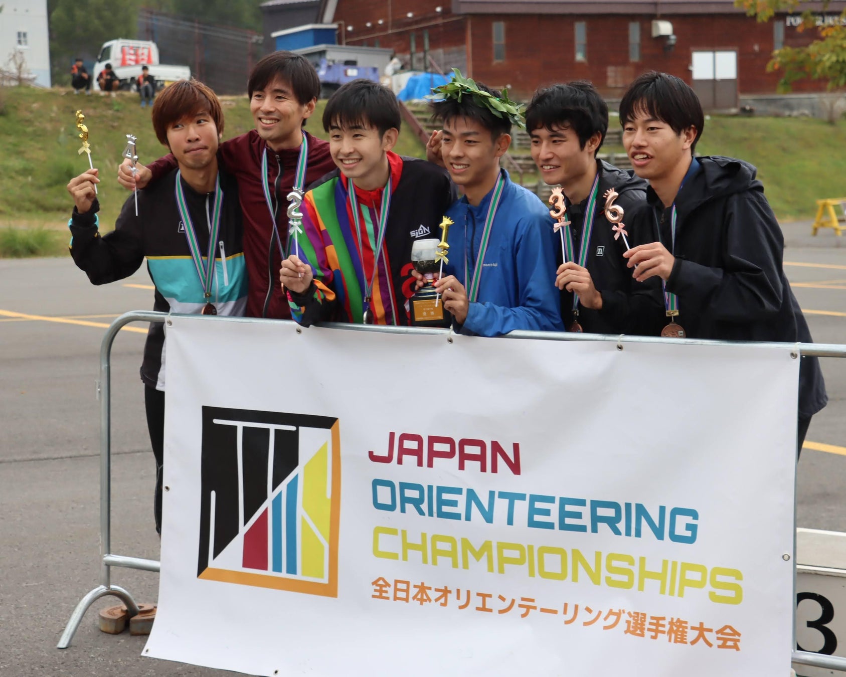 全日本オリエンテーリング選手権大会の横断幕を前に、メダルやスティックなどを手にした複数の競技者が並んで立つ記念写真。