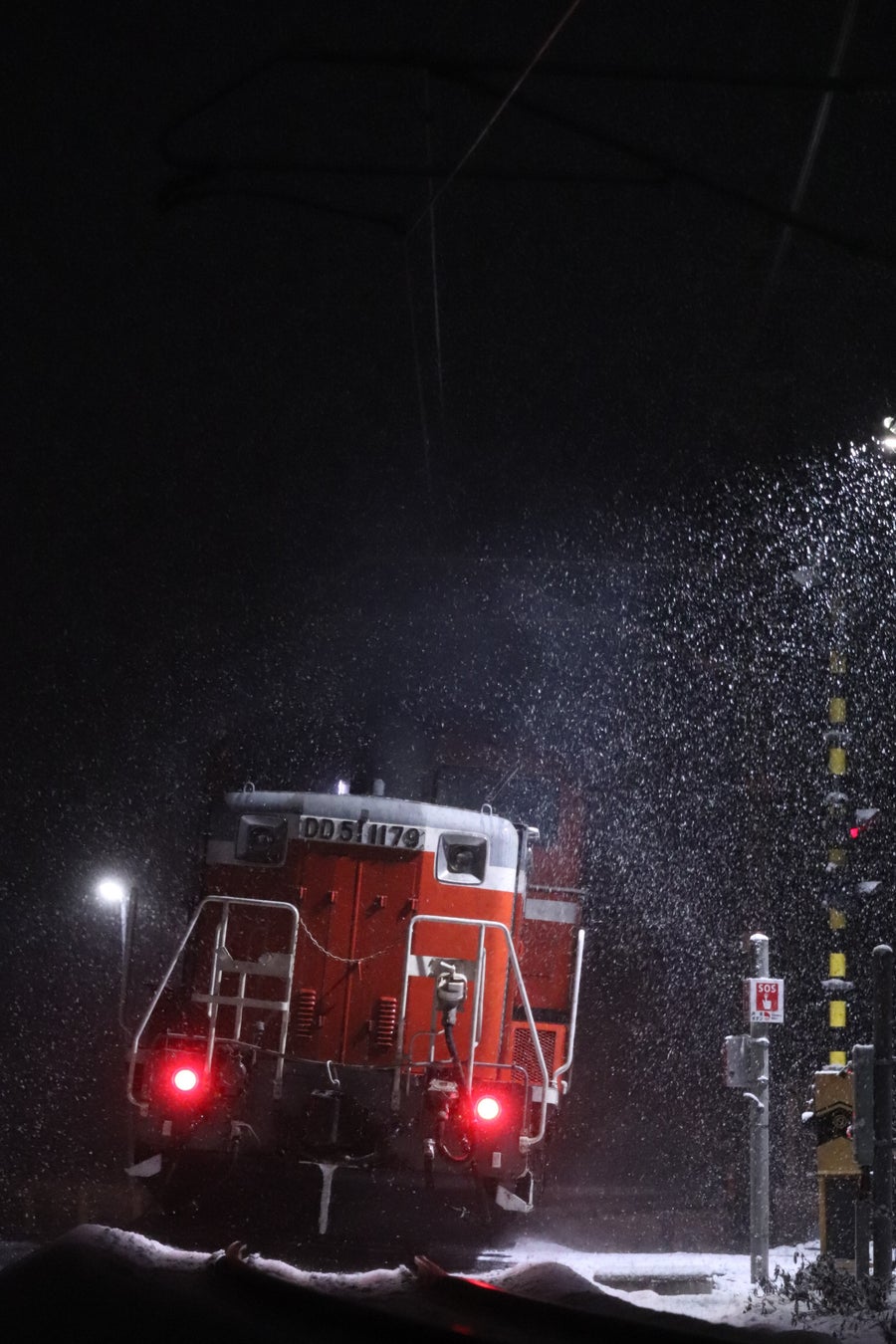 降雪の夜間に踏切の手前を照らしながら進む赤いディーゼル機関車を正面から捉えた縦構図の写真