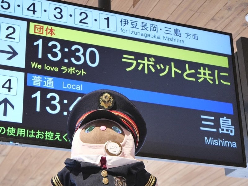 駅の電光掲示板の前に立つ制服姿のロボットの写真