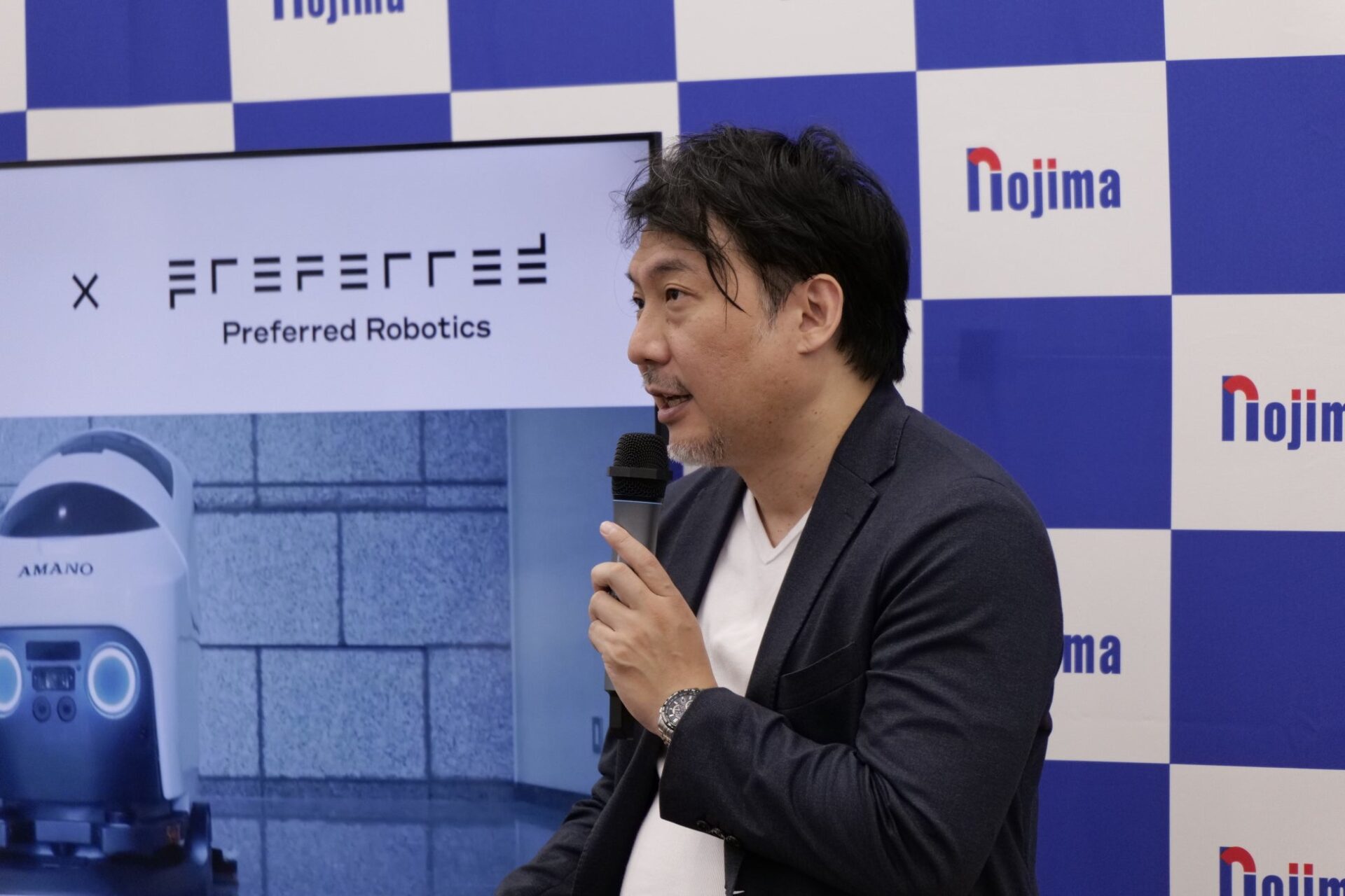 スクリーンを背景にマイクを持ち話す礒部達氏の姿