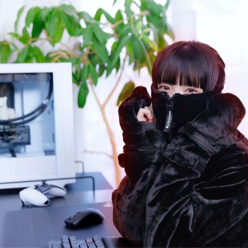 黒いウェアのフードとミトンを着用してデスクのPC前に座る人物の写真