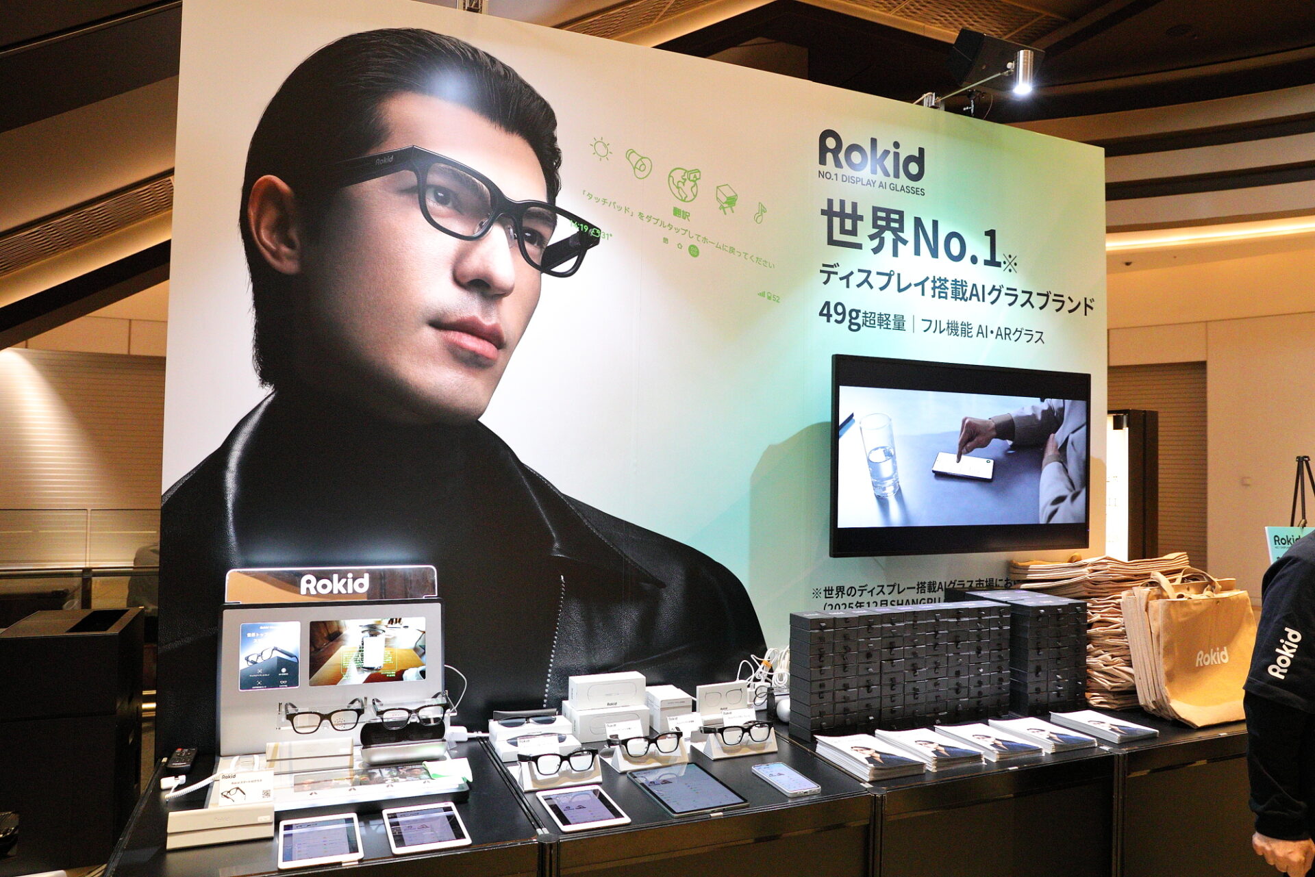 Rokidブースの展示風景。壁面のRokid広告パネルと大型モニターがあり、手前のテーブルにAIグラスやアクセサリーが並んでいる
